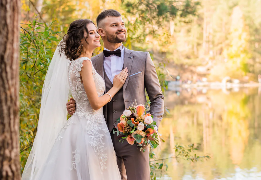 Smiling newly married couple standing by a lake in autumn light